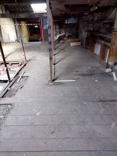 Vue intérieure d'un espace industriel délabré, au sol en bois, avec poutres apparentes et infrastructures exposées.