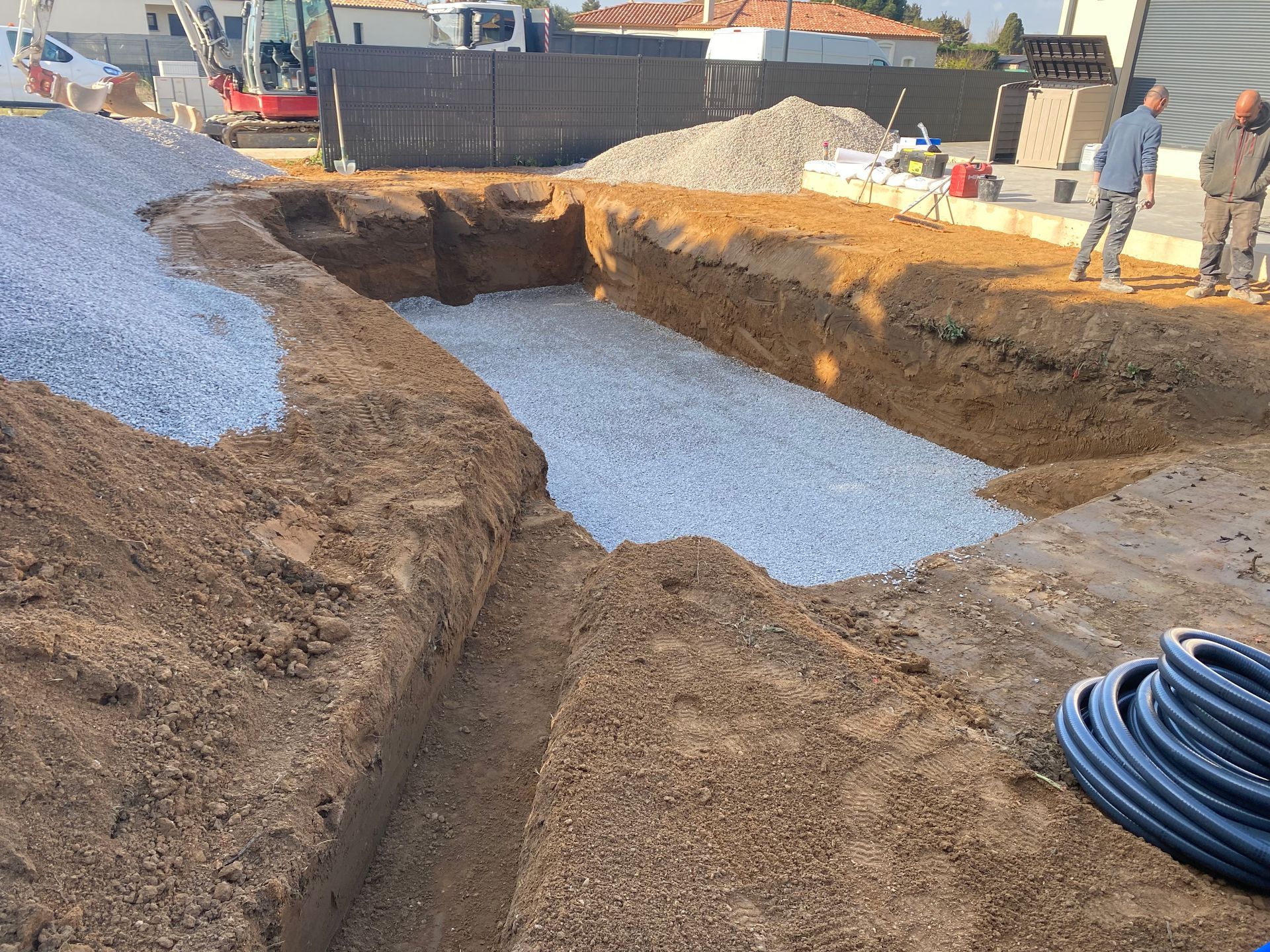 Chantier : Zone rectangulaire excavée et remplie de gravier, deux hommes se tenant à proximité.