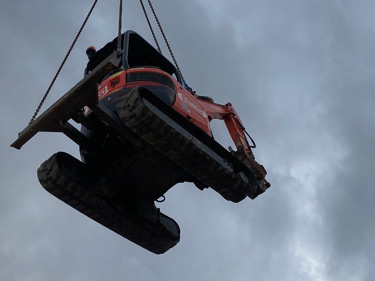 Une pelleteuse orange suspendue dans les airs par des chaînes, sur fond de ciel nuageux.