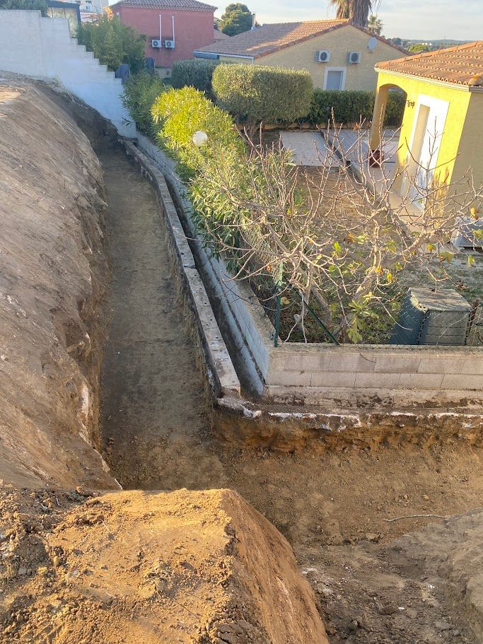 Chantier de construction avec une longue et étroite tranchée et un muret de soutènement ; bâtiments résidentiels en arrière-plan.