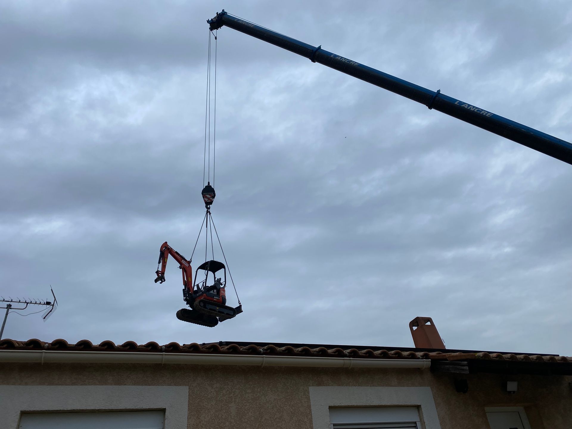 Une mini-pelle suspendue dans les airs par une grue, près d'un bâtiment sur fond de ciel nuageux.