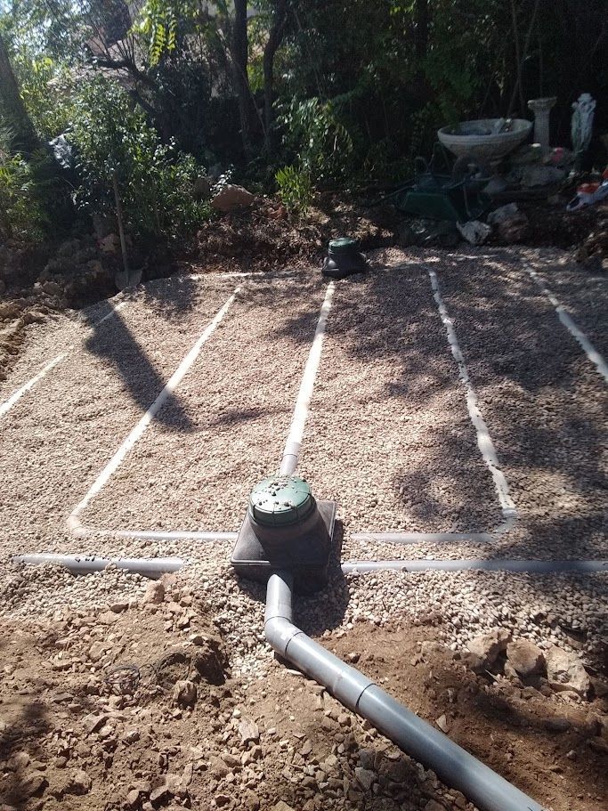 Un système septique artificiel. Des tranchées remplies de gravier avec un regard de répartition central et des tuyaux de raccordement dans une cour en terre battue.