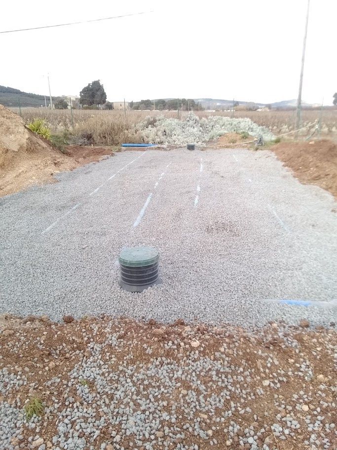Zone recouverte de gravier avec une trappe d'accès circulaire, probablement pour une fosse septique. Tuyaux bleus et terre visibles. Extérieur.