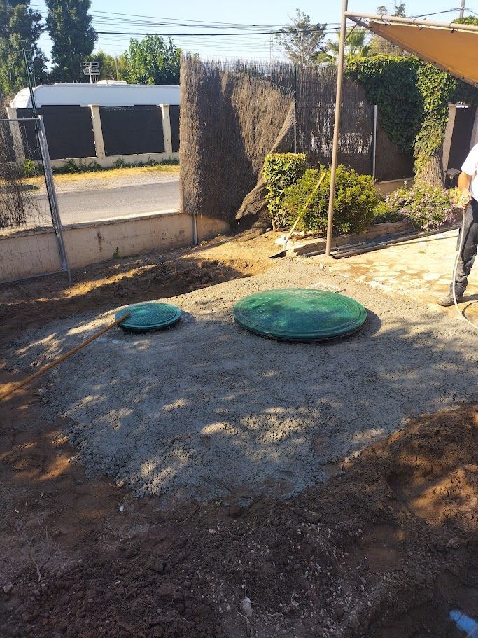 Deux couvercles circulaires verts reposent sur du gravier, recouvrant probablement des fosses septiques, à l'extérieur.