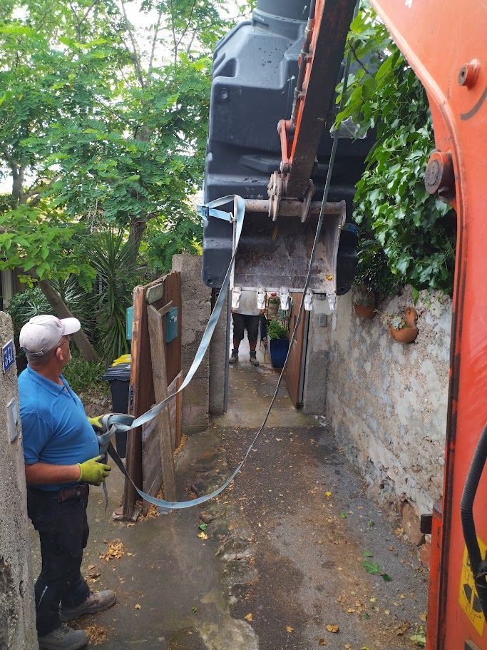 Une excavatrice utilise des sangles pour soulever des barrières en bois dans une ruelle étroite. Un ouvrier guide les sangles.