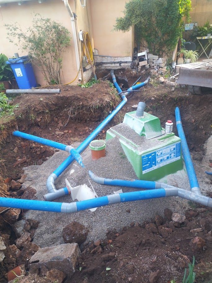 Des tuyaux bleus sont reliés à une structure verte sur un chantier de construction avec du gravier, de la terre et une poubelle bleue.