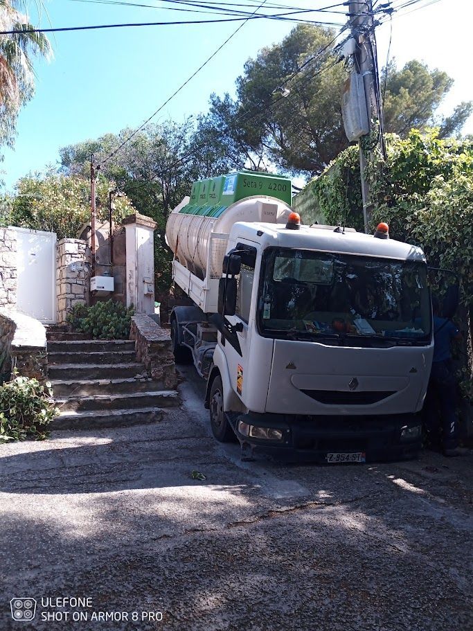 Camion transportant une citerne cylindrique stationné sur un chemin de gravier, marches et arbres visibles.