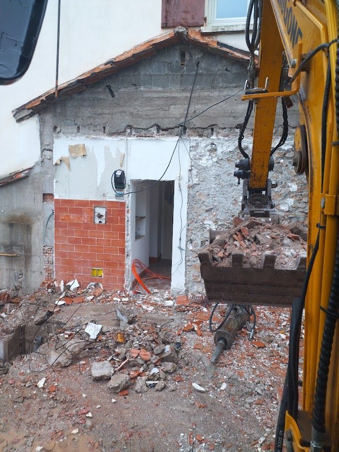 Une personne démolit un mur de briques à coups de masse, des gravats jonchent le sol, à l'intérieur.