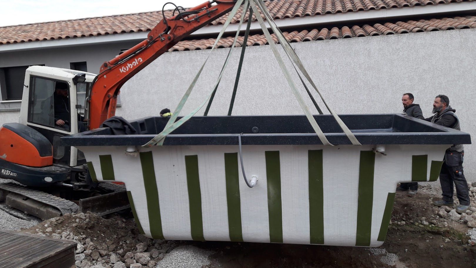 Une pelleteuse soulève la coque rectangulaire d'un grand spa, sous le regard des ouvriers. Parois à rayures vertes et blanches. En extérieur.