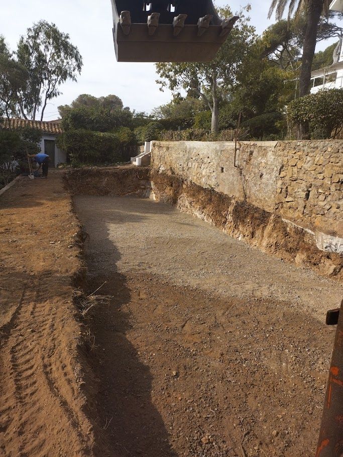 Une pelleteuse creuse une zone rectangulaire, probablement pour une piscine, près d'un mur de pierre et de végétation.