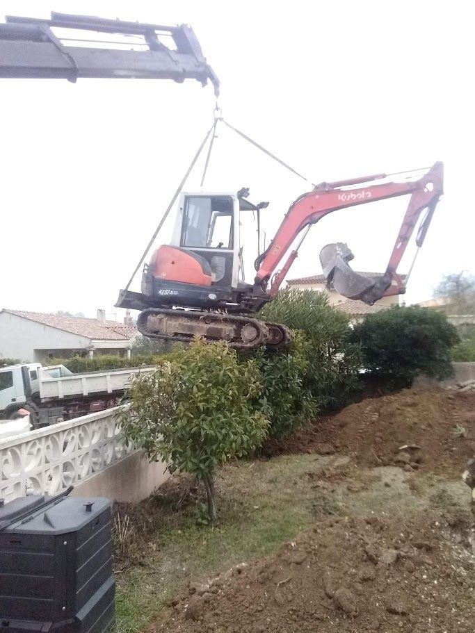 Une mini-pelle suspendue par une grue au-dessus d'une clôture, à côté d'un tas de terre et de buissons.