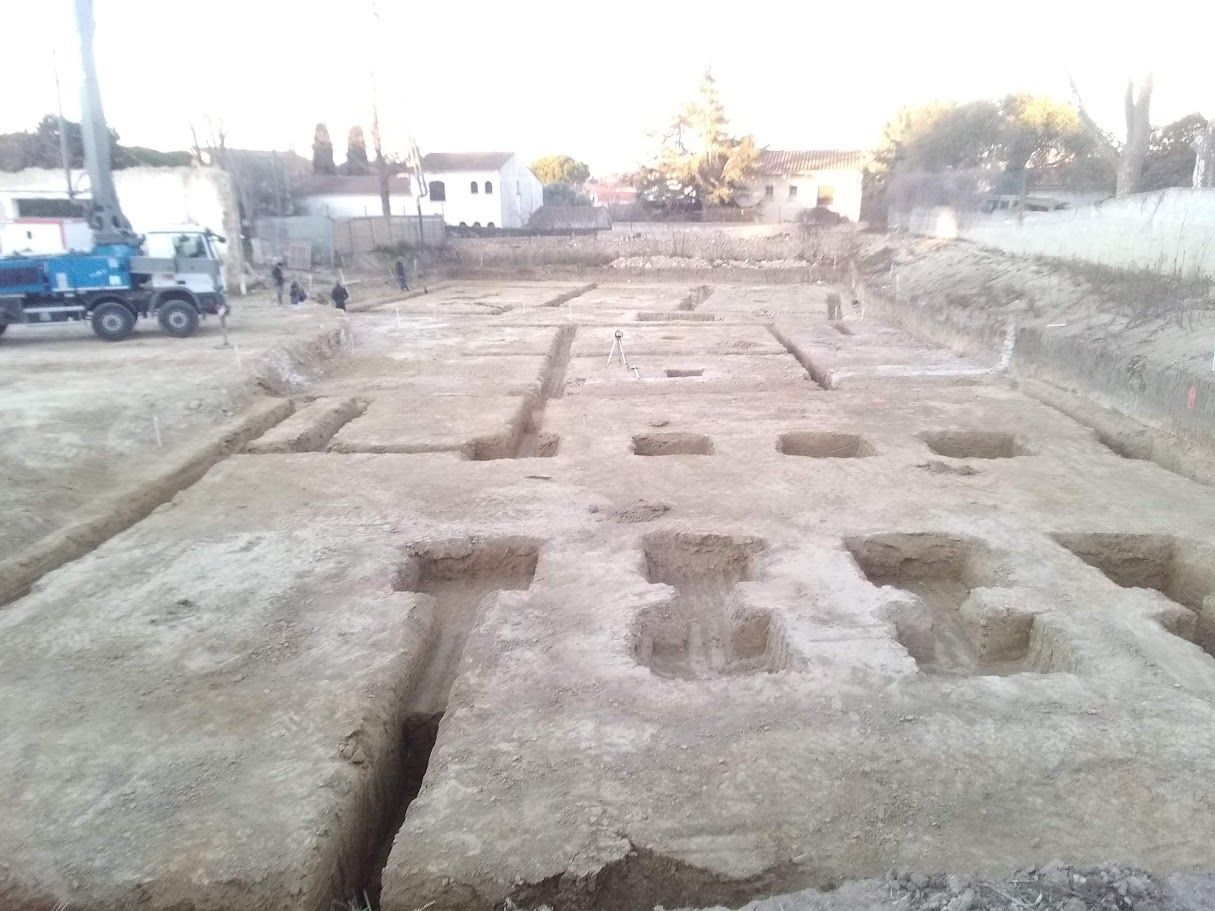 Chantier de construction avec des tranchées et des trous carrés creusés dans la terre. Un camion bleu est garé à proximité.