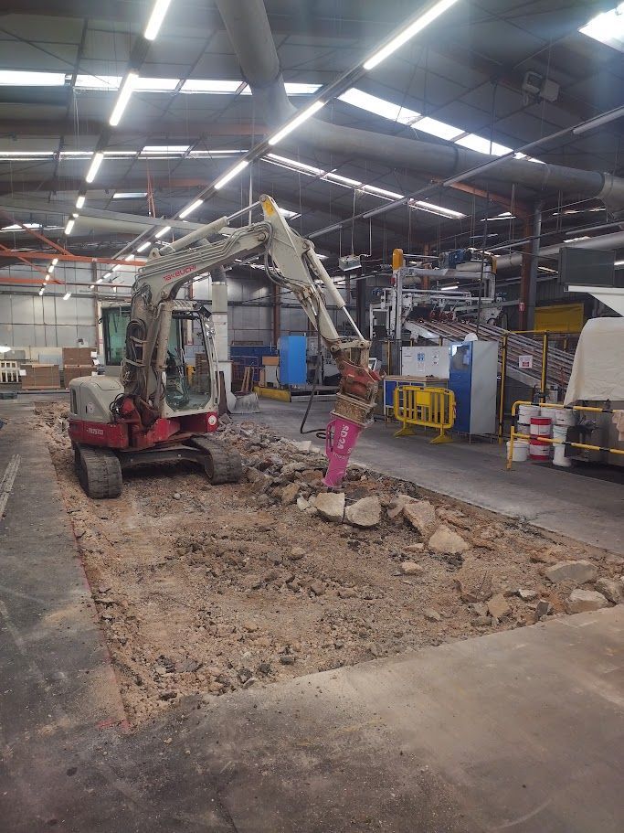 Une personne en pantalon orange creuse une tranchée à la pelle, avec une excavatrice en arrière-plan.