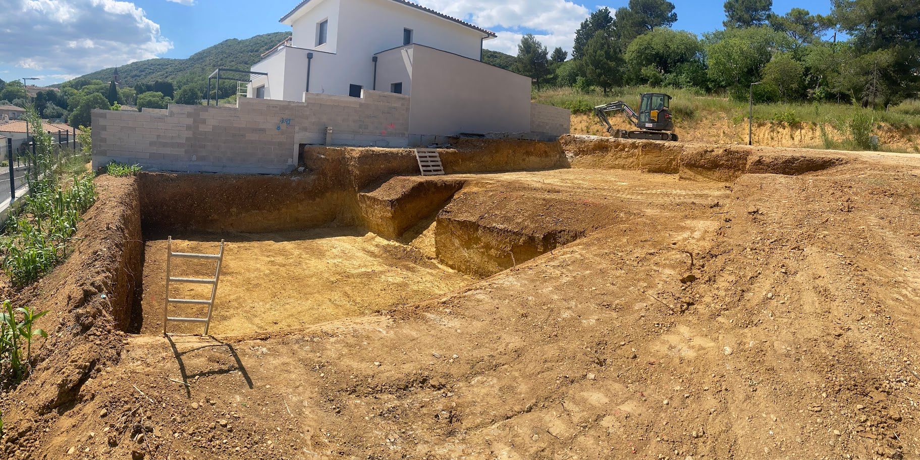 Une pelleteuse jaune creuse la terre près d'une maison par une journée ensoleillée.
