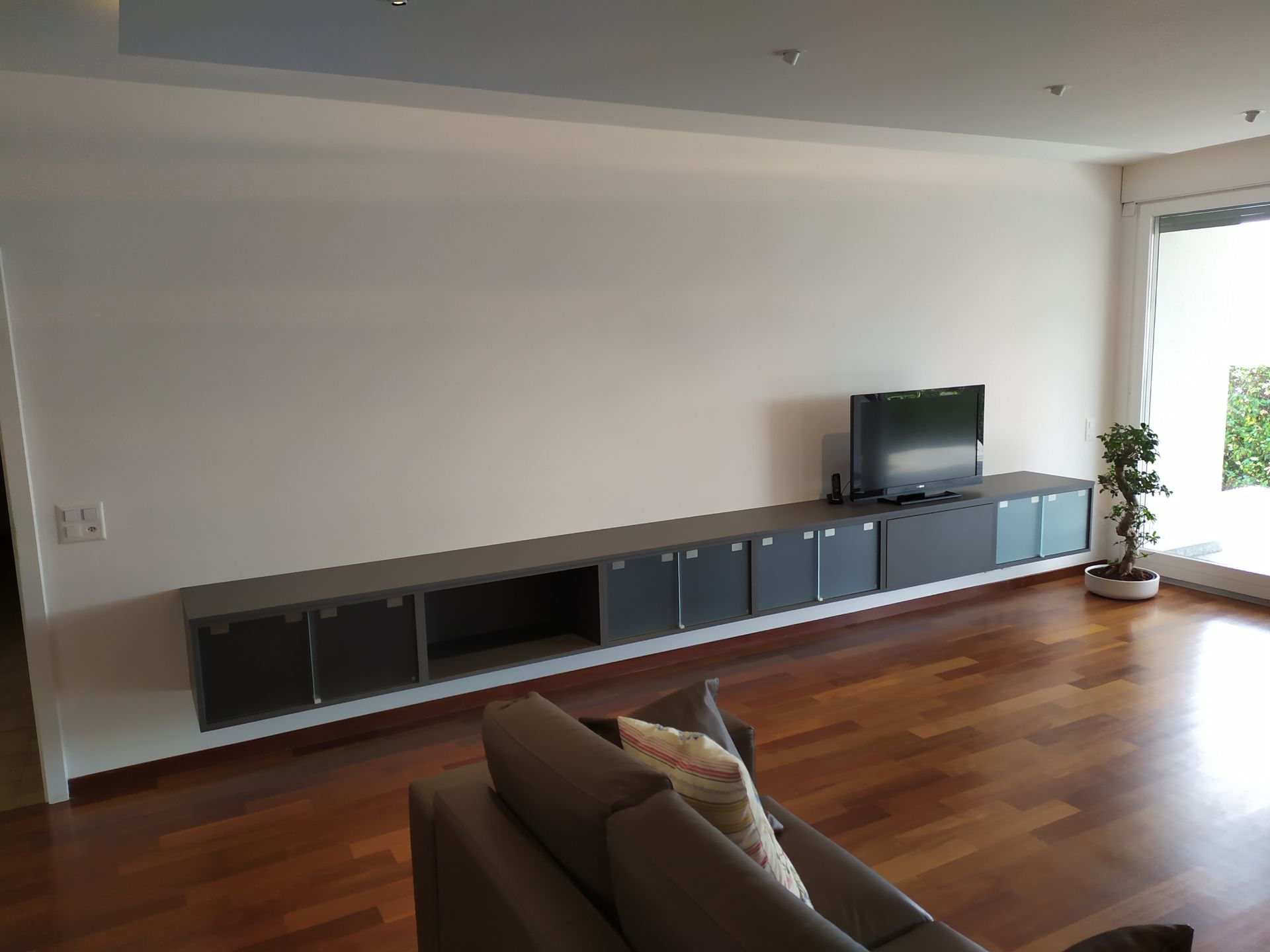 Arredamento per la sala. Esecuzione su misura. Colore grigio antracite. Vetri di sicurezza satinati bianchi. Piani di lavoro. Fissaggio alle pareti con attaccaglie nascoste e regolabili. 5 elementi con doppie antine scorrevoli in vetro. 1 elemento a ribalta per TV. 1 elemento a vista.