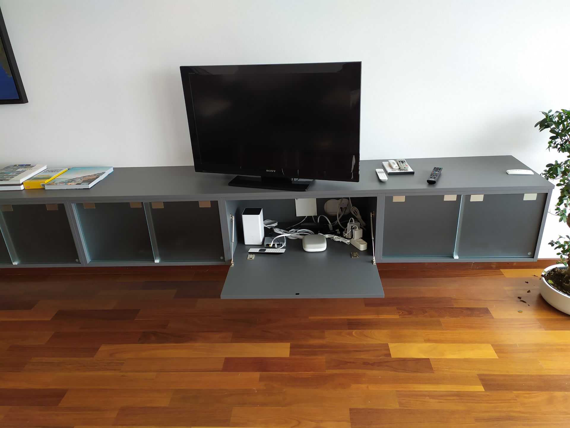 Arredamento per la sala. Esecuzione su misura. Colore grigio antracite. Vetri di sicurezza satinati bianchi. Piani di lavoro. Fissaggio alle pareti con attaccaglie nascoste e regolabili. 5 elementi con doppie antine scorrevoli in vetro. 1 elemento a ribalta per TV. 1 elemento a vista.