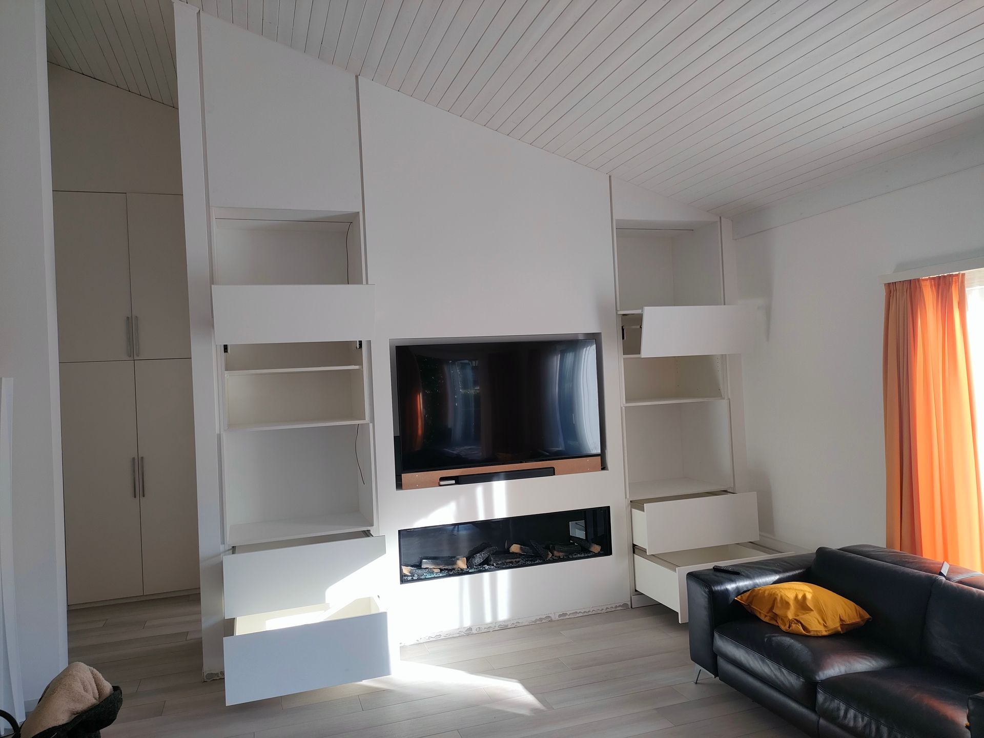 Parte centrale in cartongesso con nicchie per TV e generatore d'aria calda con animazioni realistiche e luci LED.  Parti inferiori con 2 cassettoni a estrazione totale, con blumotion e apertura a pressione. Pensili con doppie ante, impugnature nascoste, sistema di apertura BLUM Aventos HF top. Nicchie a vista con illuminazione LED a intensità regolabile. MDF grezzo pitturato come le pareti.