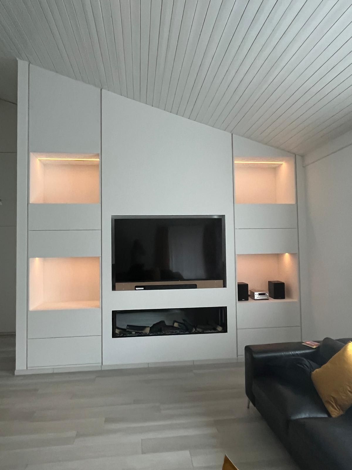 Parte centrale in cartongesso con nicchie per TV e generatore d'aria calda con animazioni realistiche e luci LED.  Parti inferiori con 2 cassettoni a estrazione totale, con blumotion e apertura a pressione. Pensili con doppie ante, impugnature nascoste, sistema di apertura BLUM Aventos HF top. Nicchie a vista con illuminazione LED a intensità regolabile. MDF grezzo pitturato come le pareti.