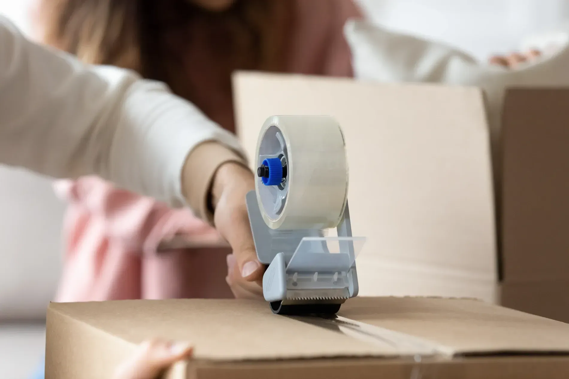Persona sellando una caja de cartón con cinta de embalaje, otros en el fondo.