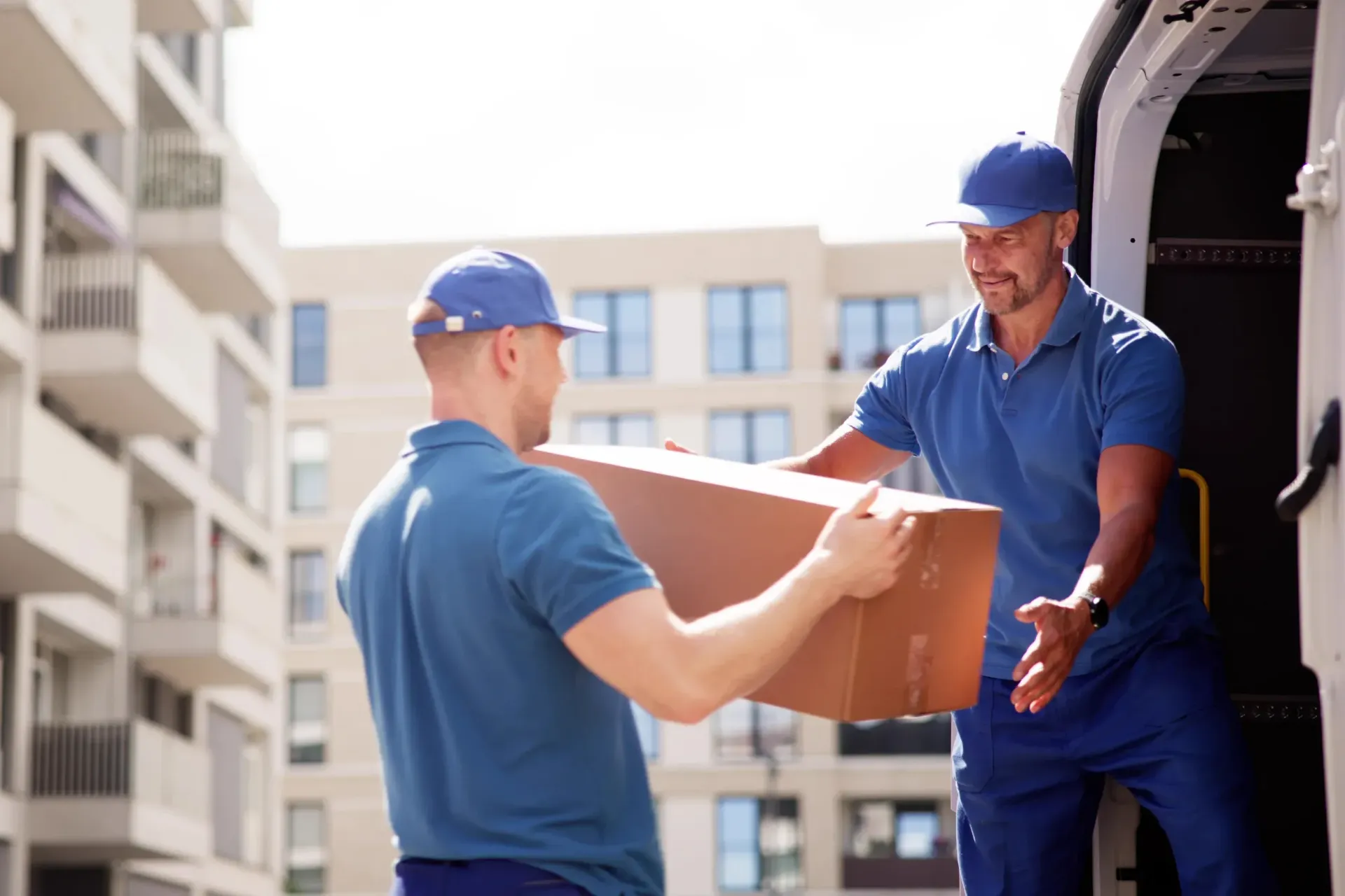 Dos trabajadores de mudanzas con monos azules llevan un sillón beige en una habitación luminosa con cajas.
