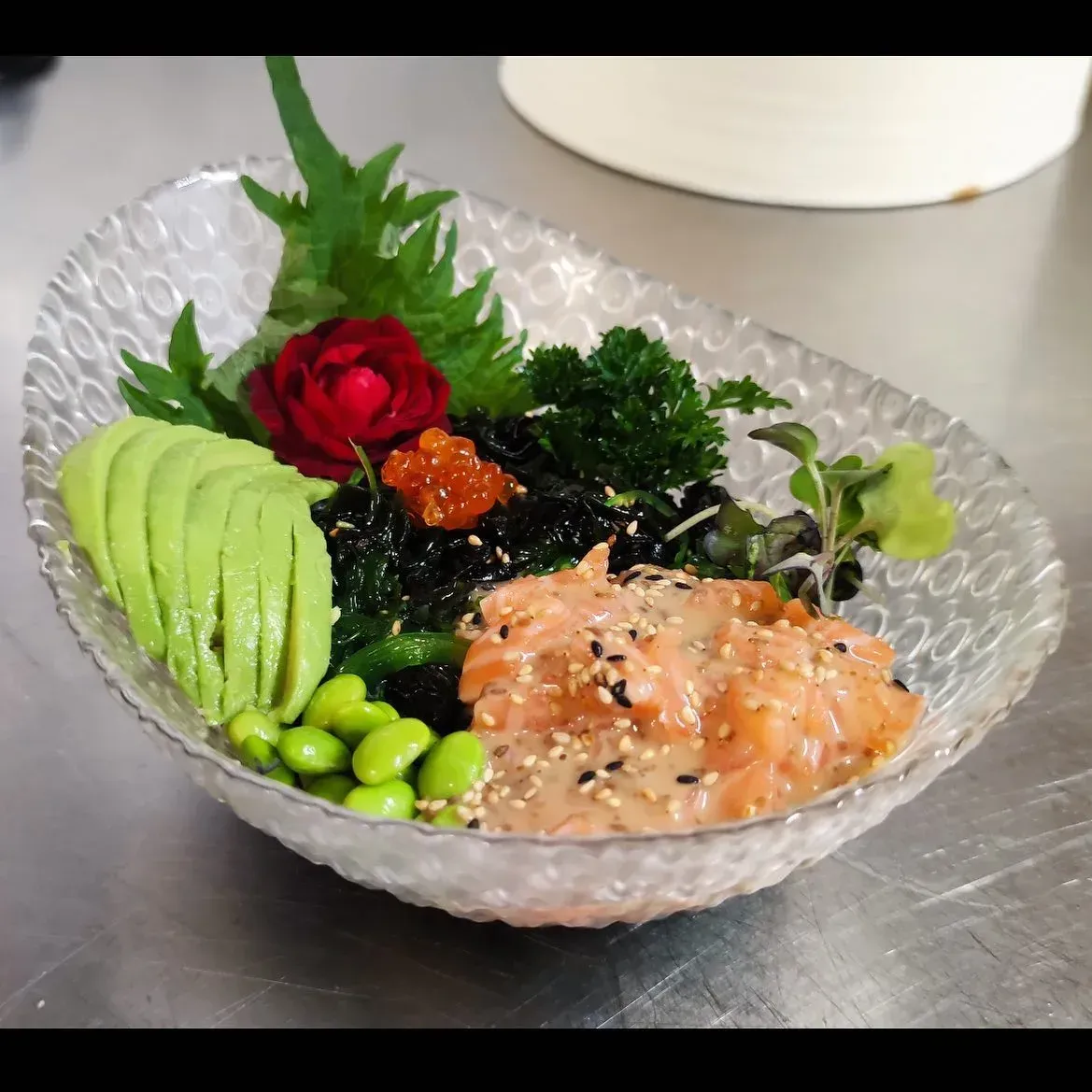 Cuenco de vidrio con sushi variado: salmón, rodajas de aguacate, edamame, algas y guarnición sobre una encimera metálica