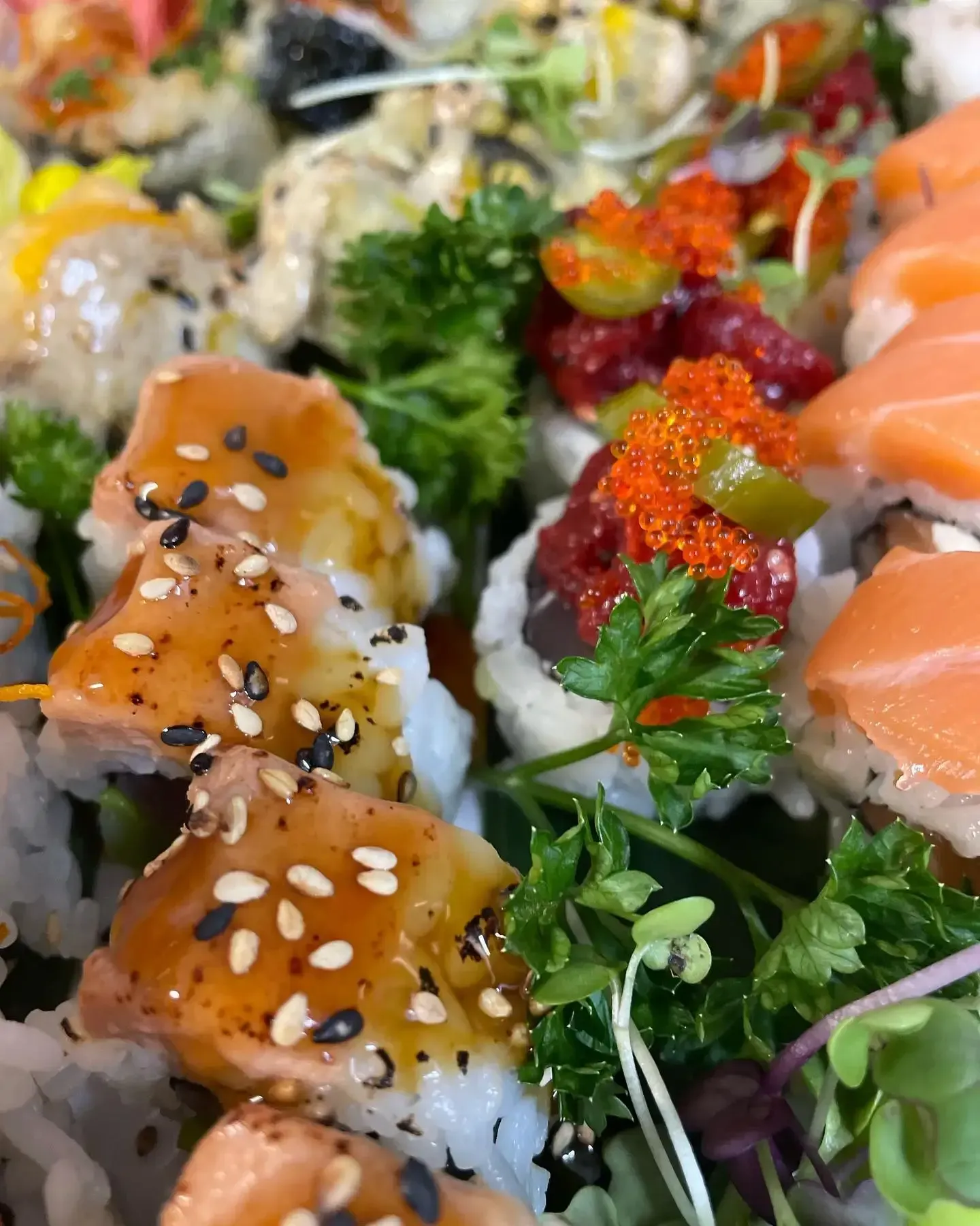 Variedad de rollos de sushi cubiertos con salmón, sésamo y huevas de pescado, decorados con verduras frescas.