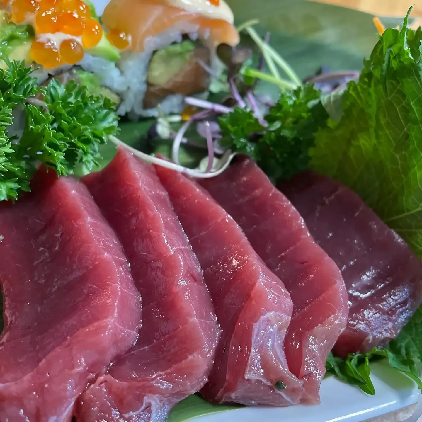Sashimi de atún crudo en lonchas, adornado con perejil y hojas verdes en un plato, con huevas de naranja y salsa por encima