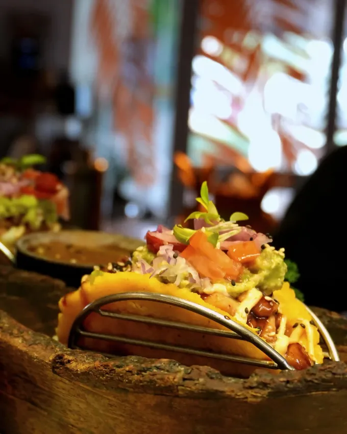 Taco en un cuenco de madera, cubierto de verduras, sobre una mesa de restaurante con el fondo desenfocado