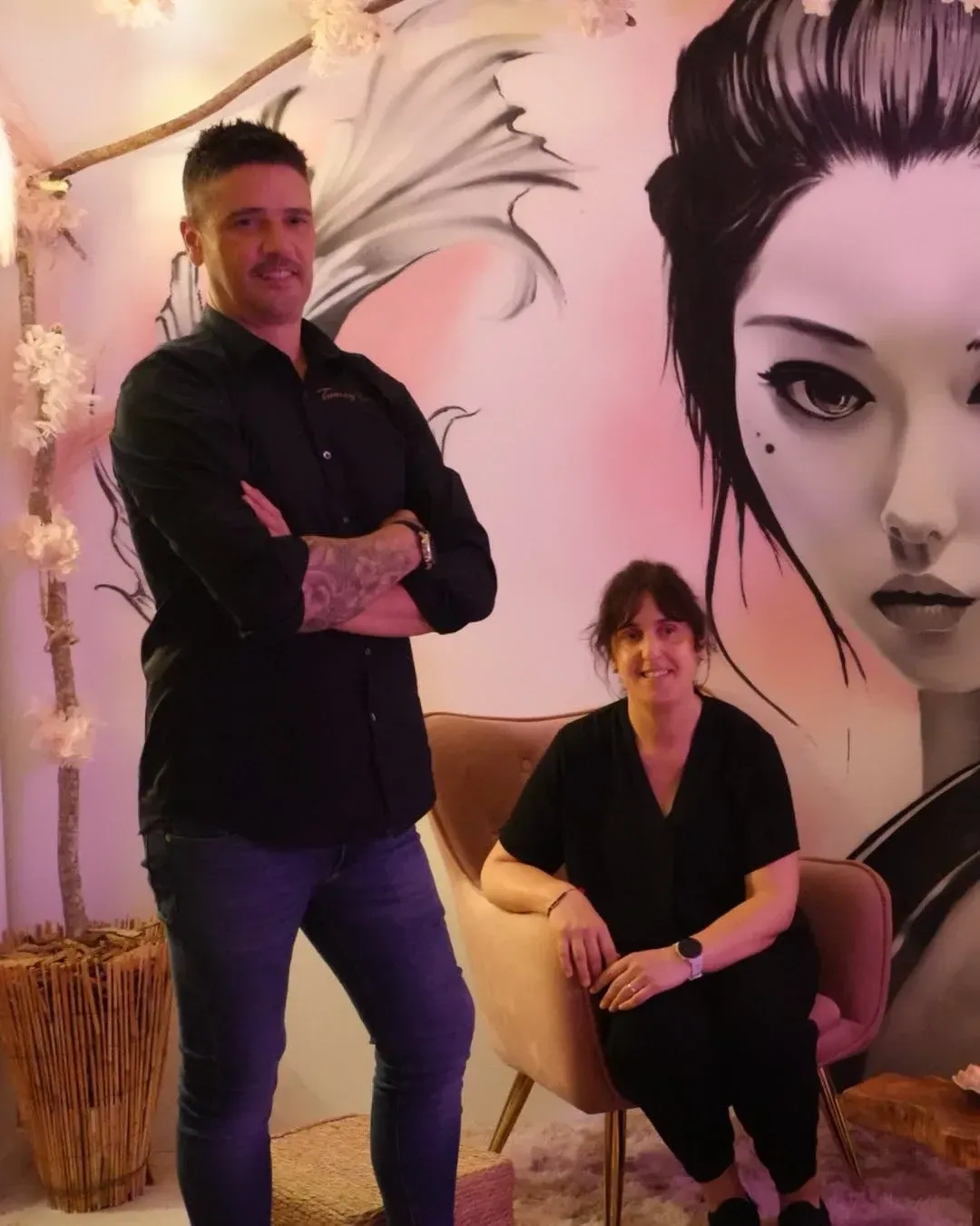 Dos personas posando en una habitación iluminada de rosa, con un gran mural del rostro de una mujer en la pared
