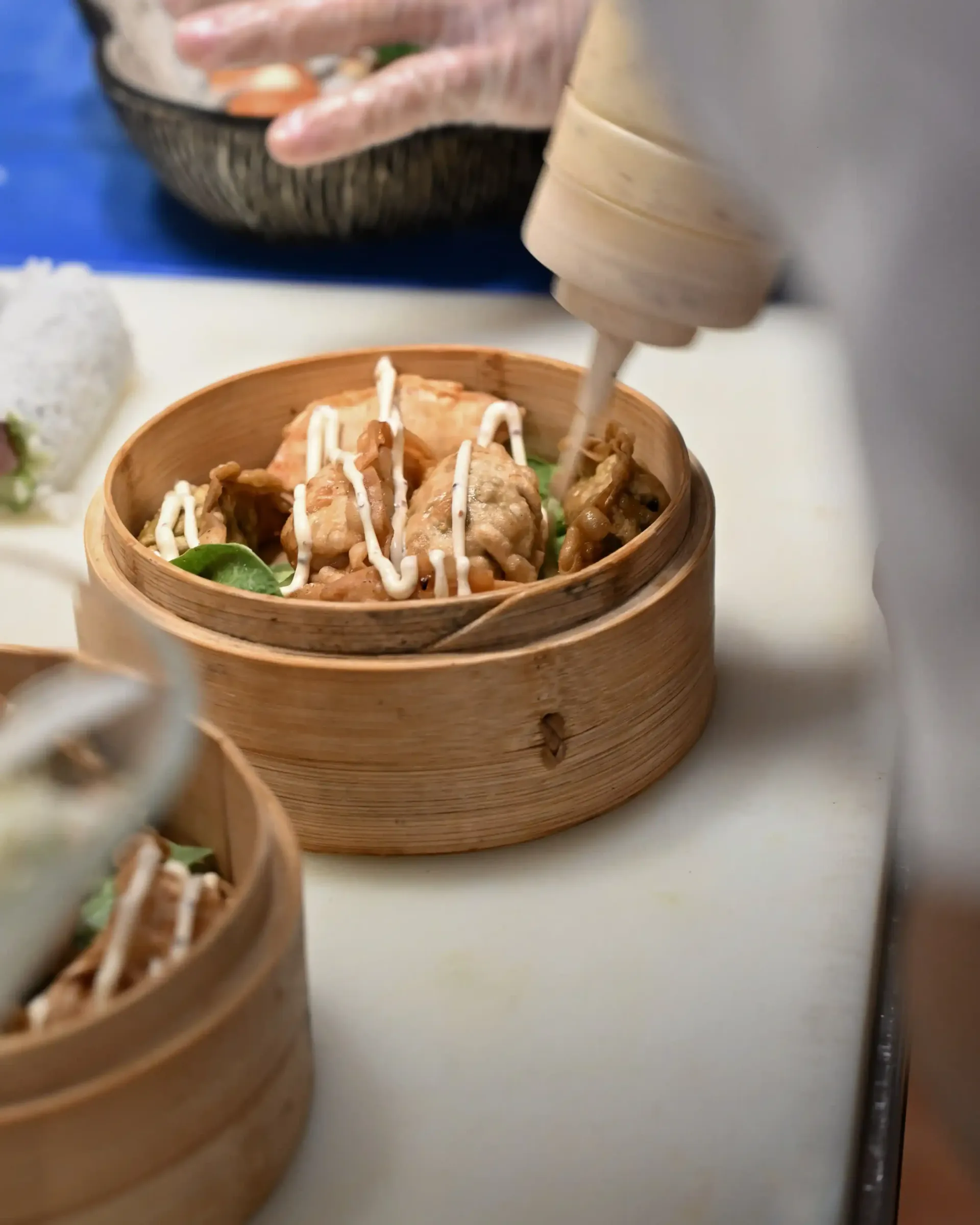 Una cesta de dumplings al vapor con salsa rociada por encima, sobre la encimera de preparación de un restaurante