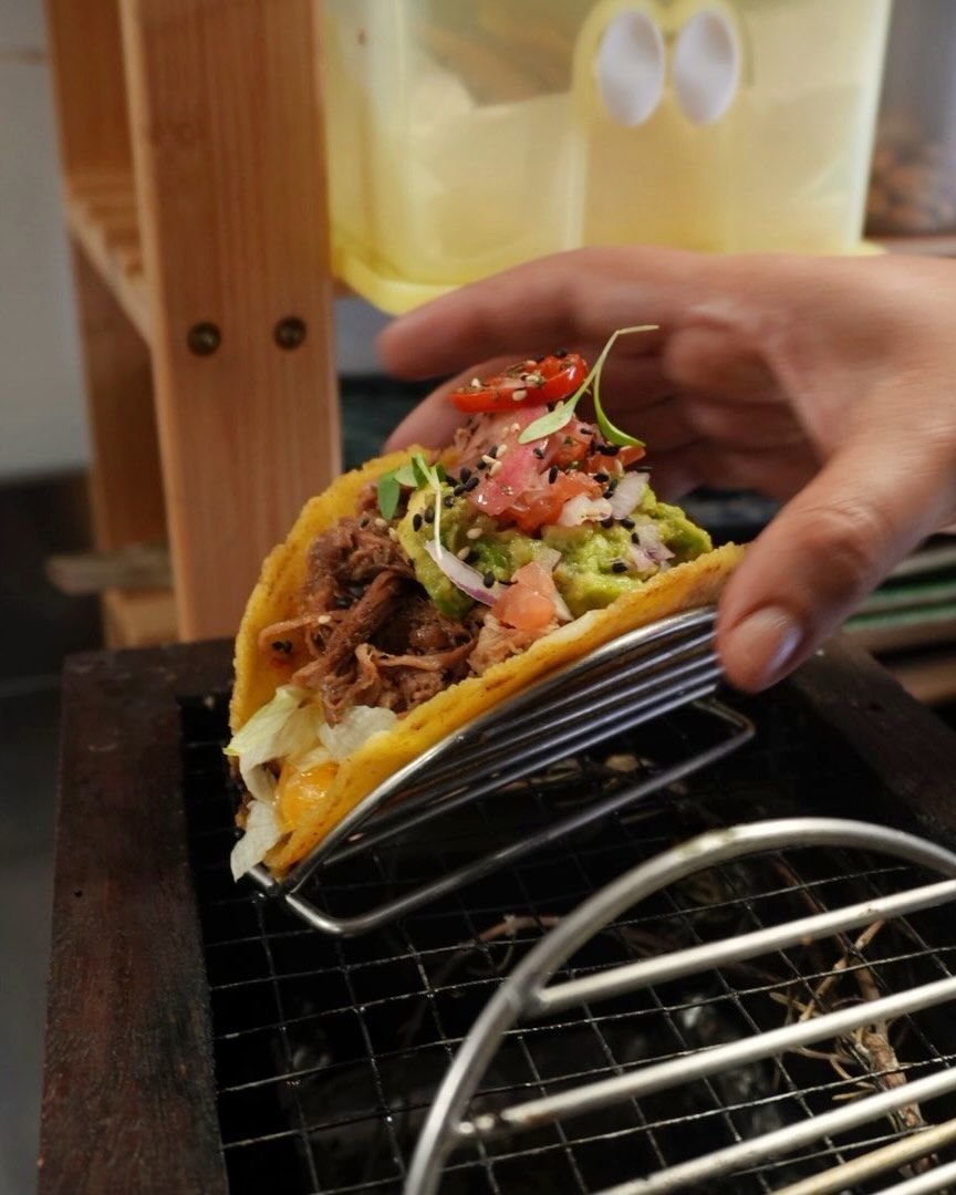 Mano sosteniendo un taco de carne deshebrada, queso y salsa sobre una rejilla metálica en un ambiente informal