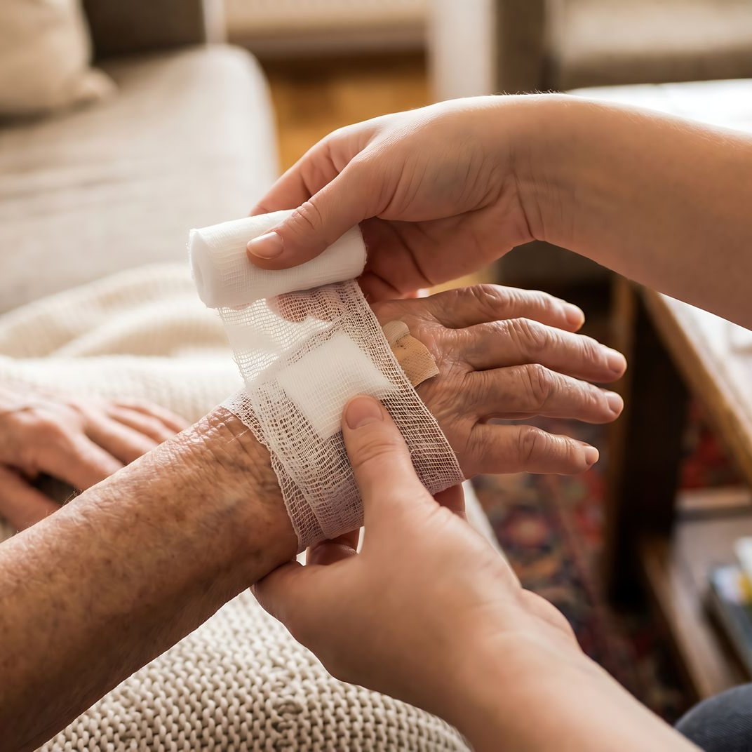 Des mains posent un bandage autour du poignet d’une personne âgée assise sur un canapé.
