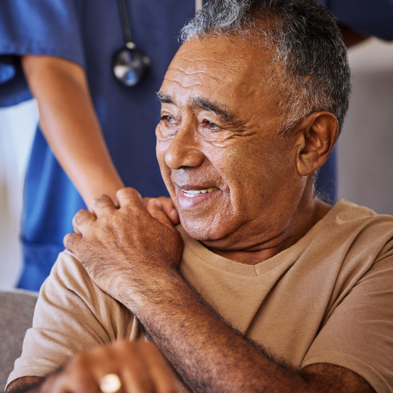 Un homme souriant est assis sur un canapé tandis qu’un soignant en bleu se tient derrière lui avec un stéthoscope.