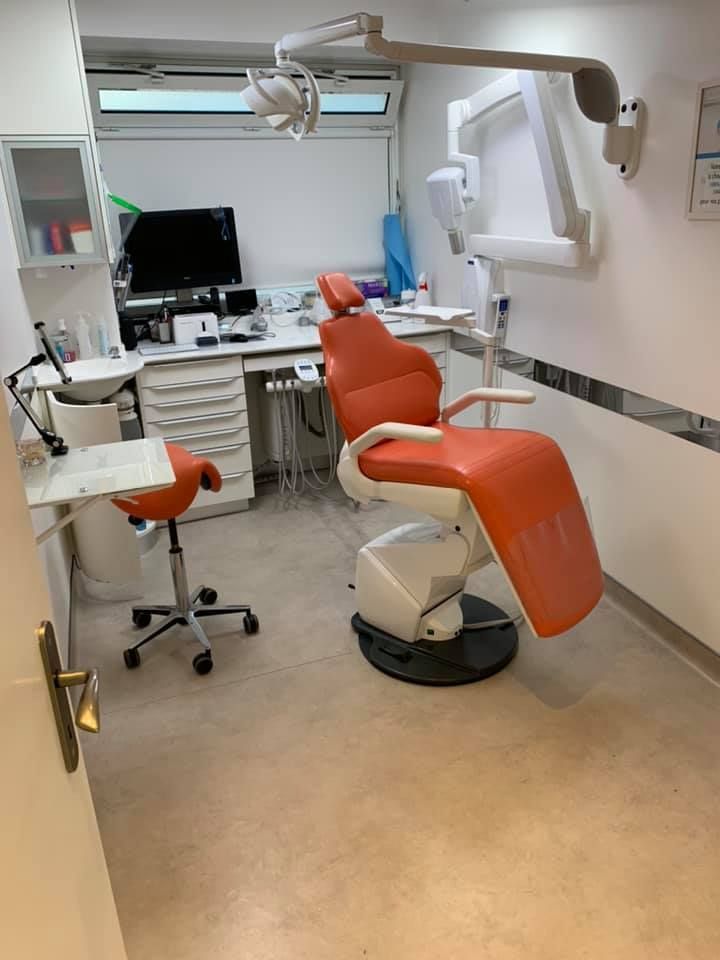 Cabinet dentaire avec chaise orange, tabouret à roulettes, bureau, ordinateur et lampe murale.