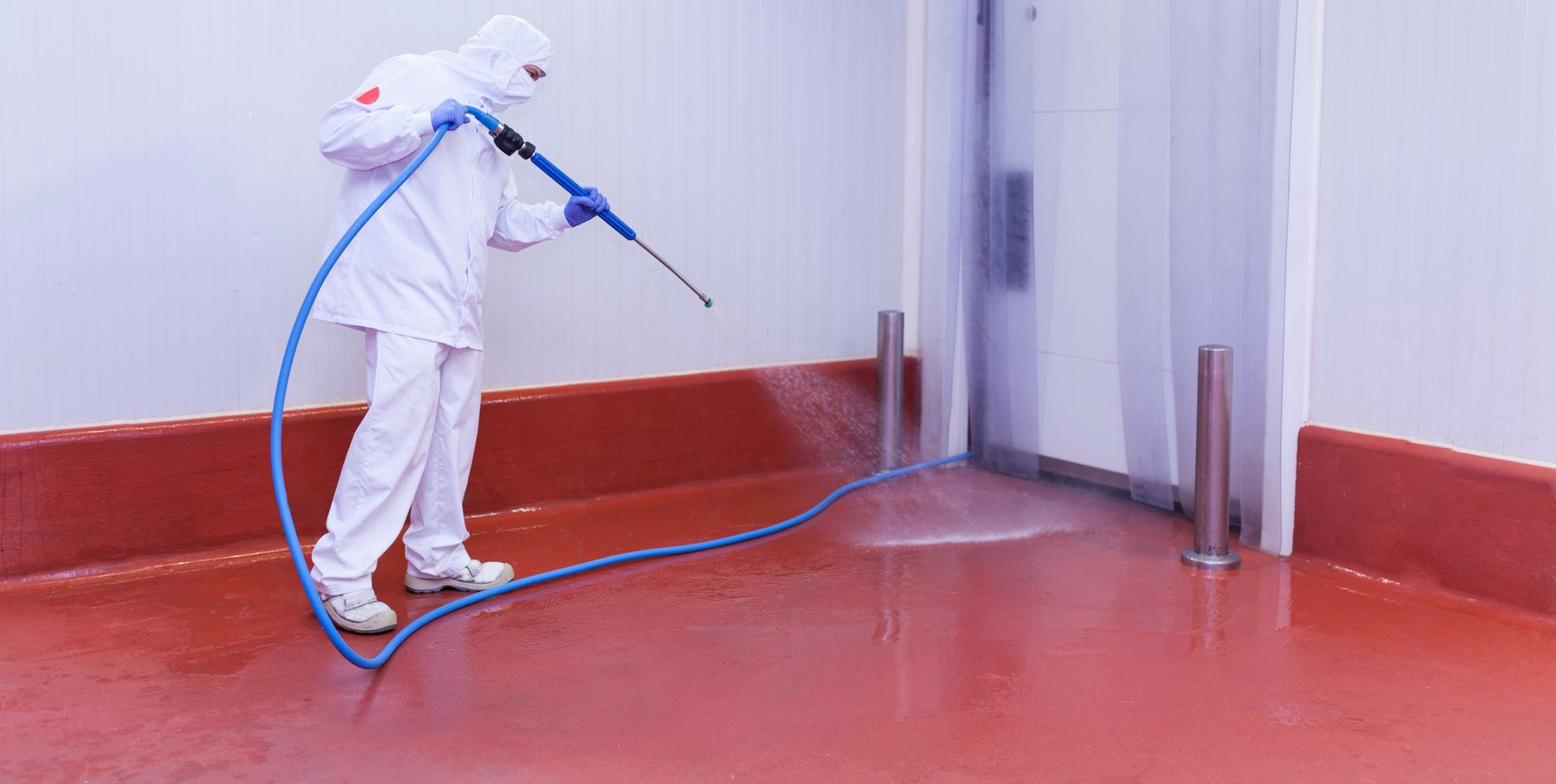 Dans une installation industrielle, une personne vêtue d'une combinaison de protection blanche nettoie un sol au jet d'eau.