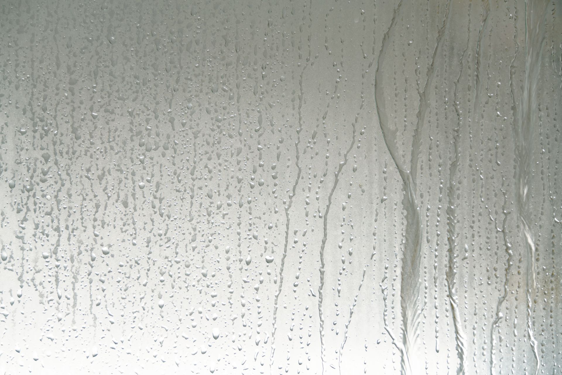 Des gouttes de pluie et des traînées d'eau ruissellent sur une surface de verre lisse et grise.