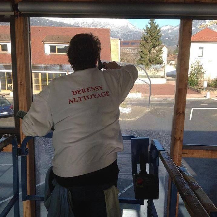 Une personne portant un sweat-shirt blanc Derensy Nettoyage utilise une raclette pour nettoyer une vitre depuis une nacelle.