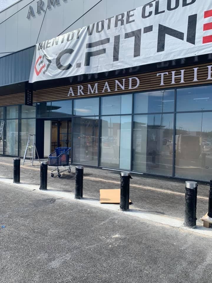 Façade extérieure avec grandes vitrines, une banderole et une rangée de bornes noires au premier plan.