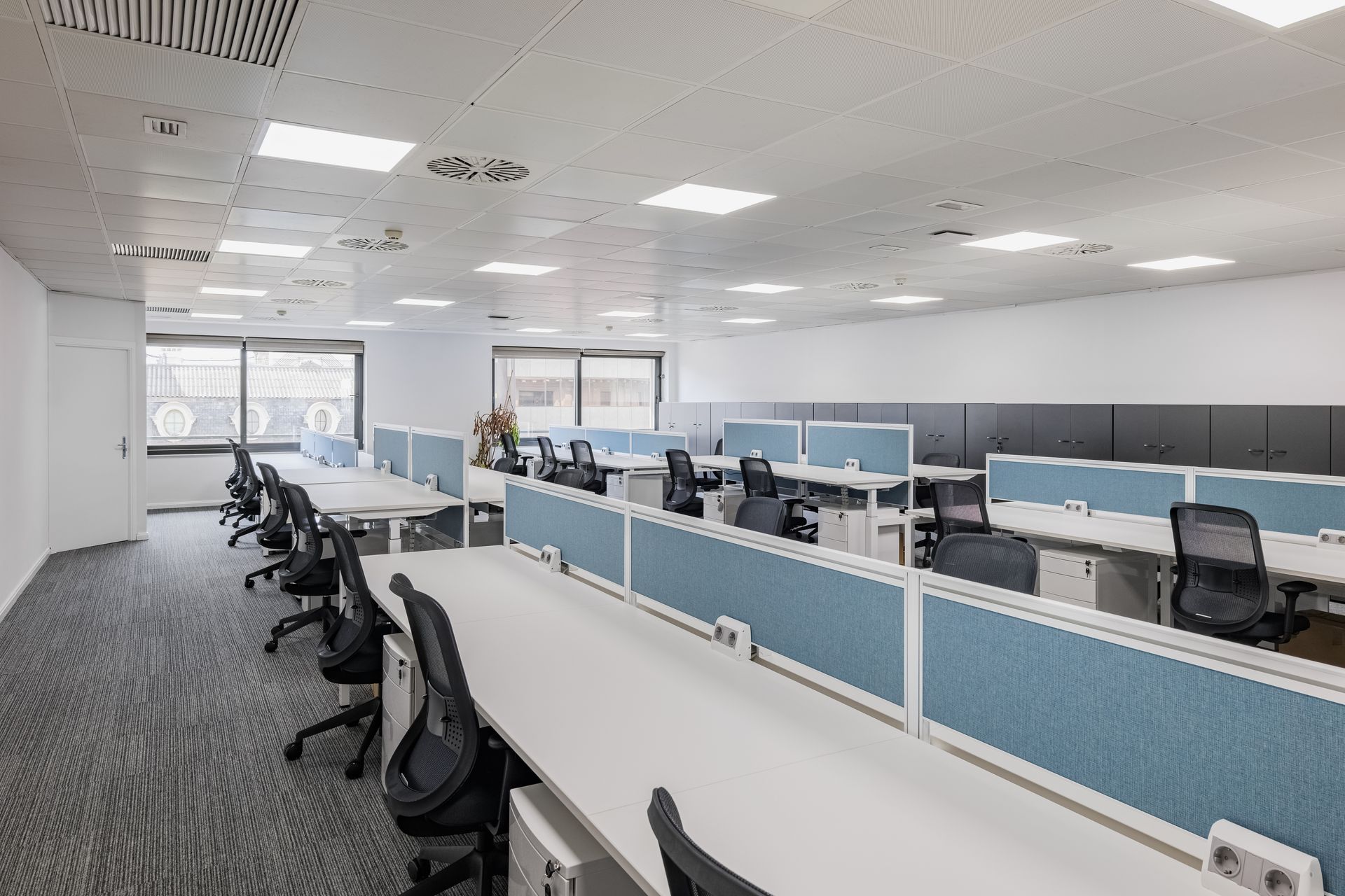 Openspace moderne avec des rangées de bureaux, des cloisons et des chaises à roulettes sur une moquette.