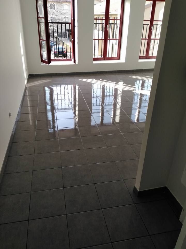 Une pièce vide avec un sol carrelé gris foncé poli et trois grandes fenêtres à cadre rouge donnant sur une rue.