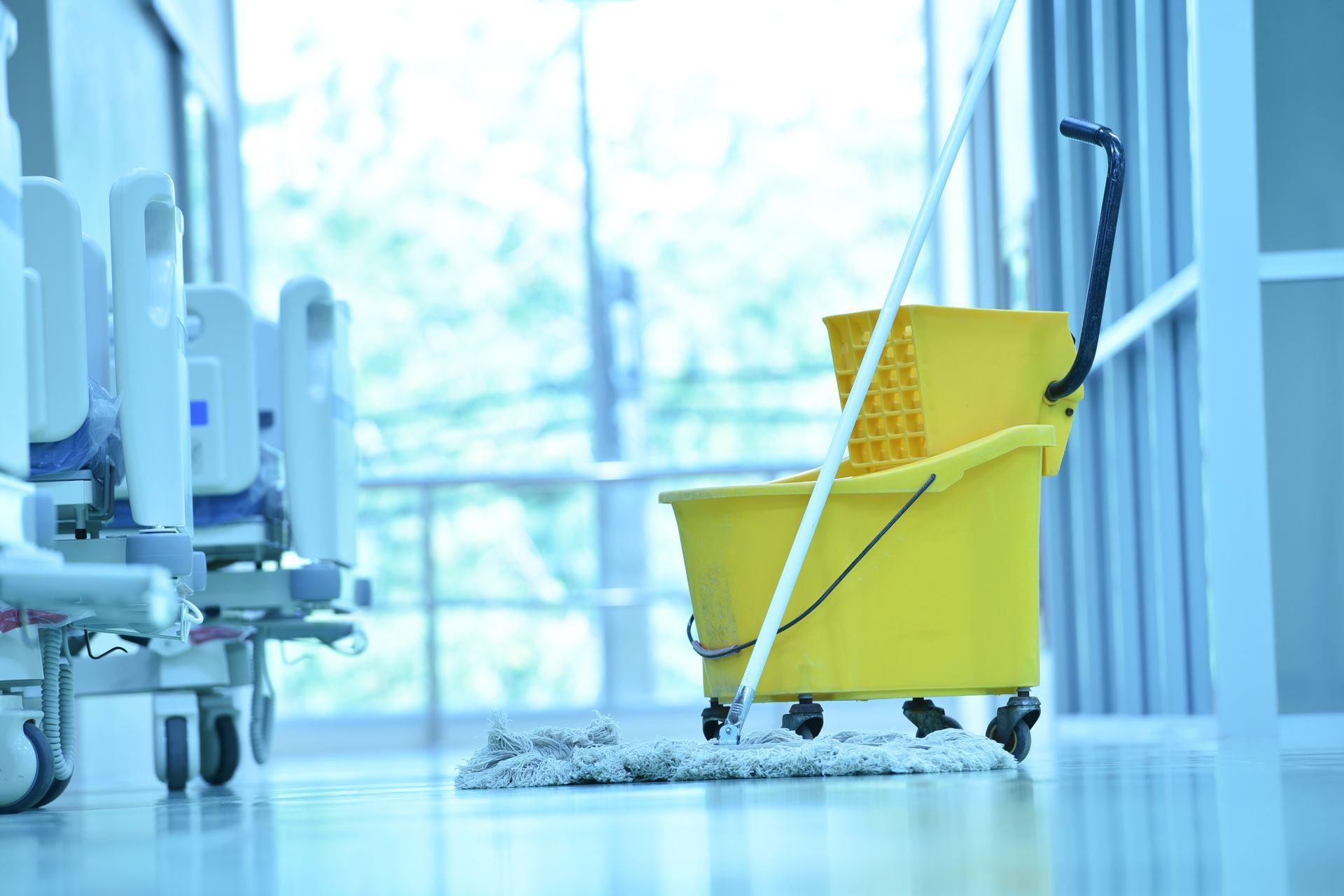 Un seau à serpillière industriel jaune sur roulettes, contenant une serpillière, est posé sur le sol d'un couloir d'hôpital.