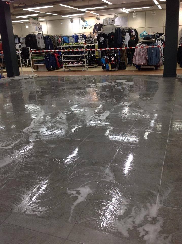 Sol carrelé d'un magasin de vêtements recouvert d'eau savonneuse, reflétant les lumières au plafond et les articles en stock.