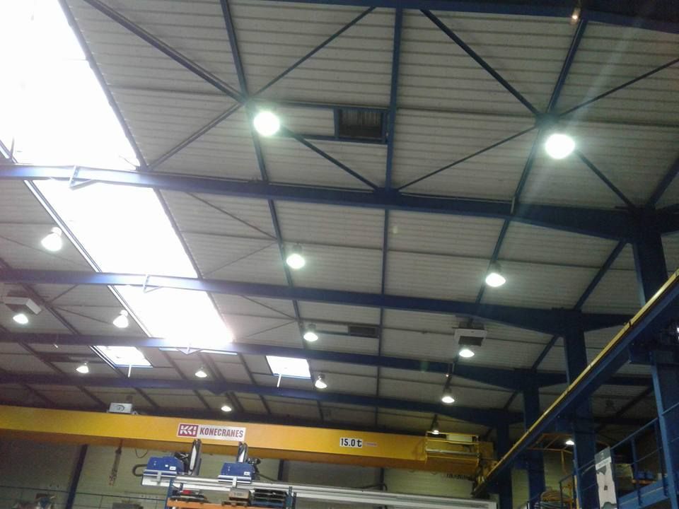 Vue en contreplongée du plafond d'un grand entrepôt avec poutres métalliques apparentes, éclairages et pont roulant.