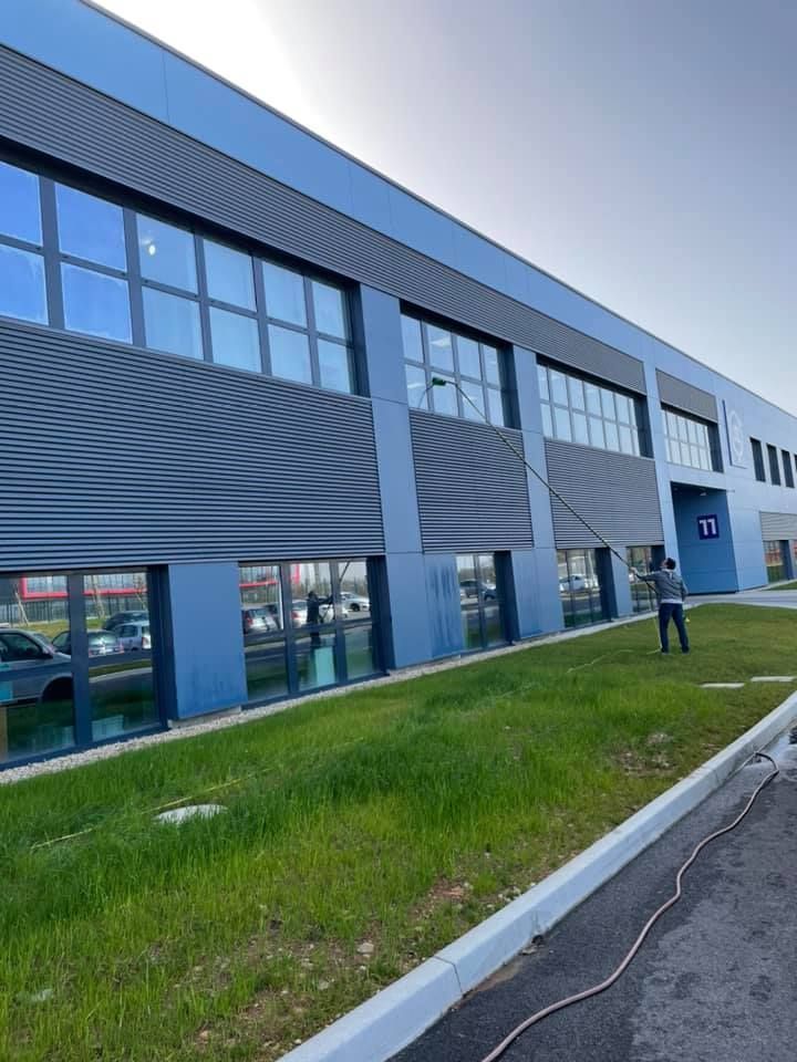 Une personne nettoie les vitres d'un bâtiment industriel à l'aide d'une perche télescopique.