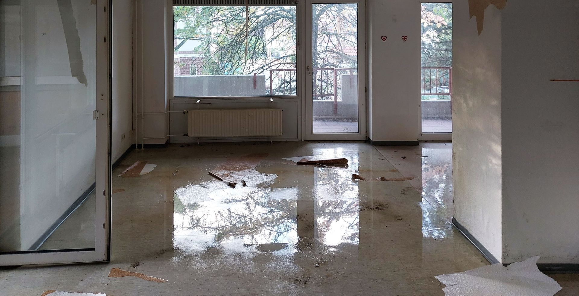 Pièce intérieure inondée avec de l'eau stagnante au sol, des débris et une vue sur l'extérieur à travers les fenêtres.