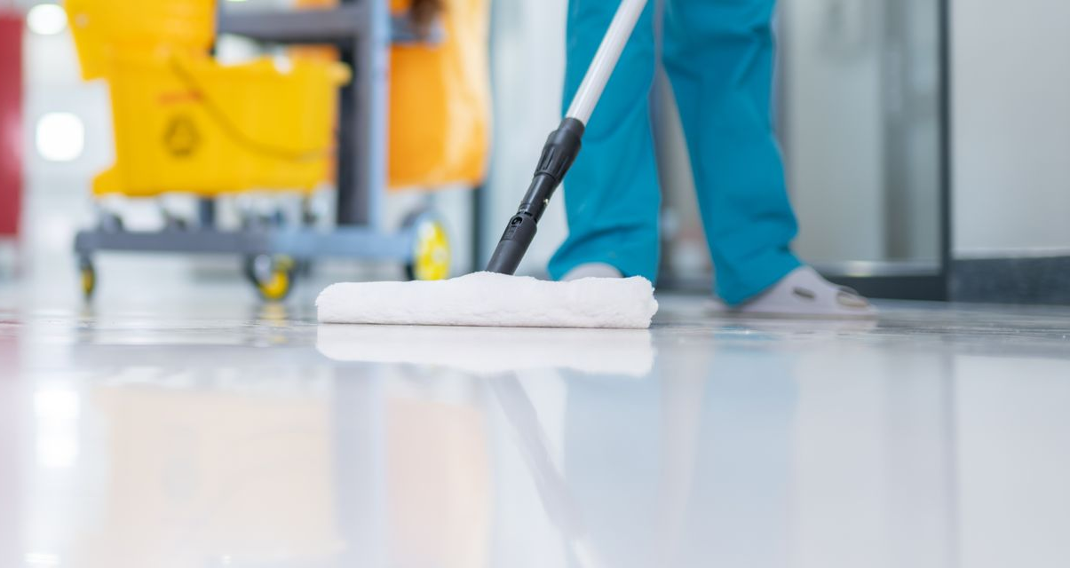 Dans un bâtiment moderne et lumineux, une personne lave un sol blanc ciré à côté d'un chariot de nettoyage jaune.
