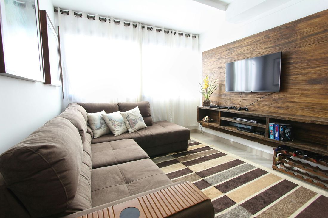 Wohnzimmer mit braunem Sofa, Fernseher auf Holzpaneel, Teppich und weißen Vorhängen.