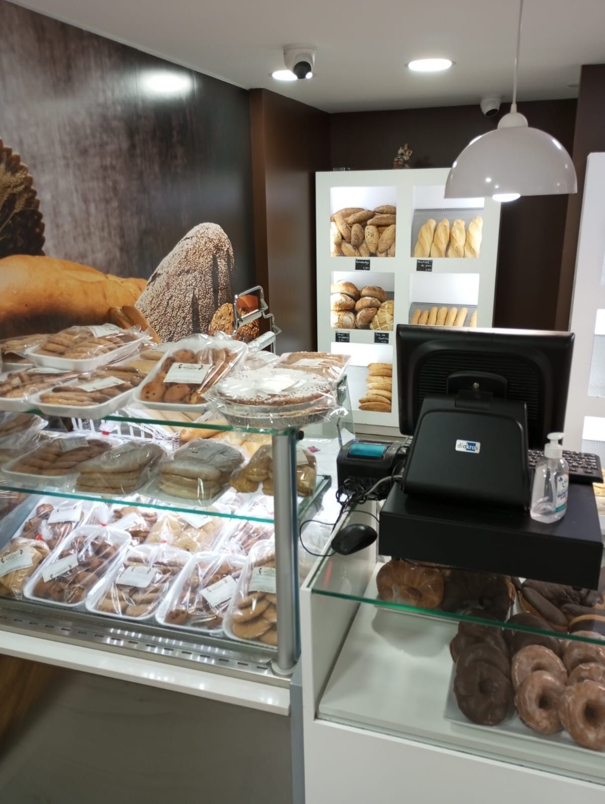 Un mostrador de panadería con vitrinas de cristal repletas de pasteles, pan y rosquillas, y un terminal de punto de venta.