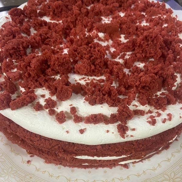 Un pastel de terciopelo rojo con glaseado de crema blanca y migas de pastel rojas por encima, sobre un plato blanco estampado.