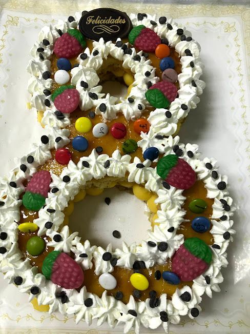 Una tarta con forma de número 8 decorada con glaseado blanco, caramelos de colores, frambuesas y un letrero que dice 